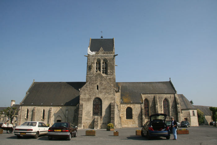 Kirken i Saint-Mere-Eglise, hvor der hænger en dukke i kirkespiret, forestillende en engelsk soldat, der var så uheldig at "lande" der. Han lod, som om han var død og overlevede på den måde. 