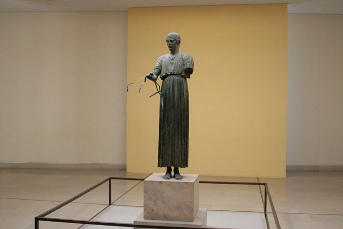 Delfi Arkæologiske Museum. Den unge vognstyrer står fastfrosset i bronze, blikket er rettet fremad, og højre hånd holder løst i tømmerne. Statuen vidner om Delfis pythiske lege.
