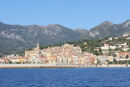 Menton set ude fra havet. Byen ligger flot placeret op ad en bjergskråning med høje bjerge i baggrunden.