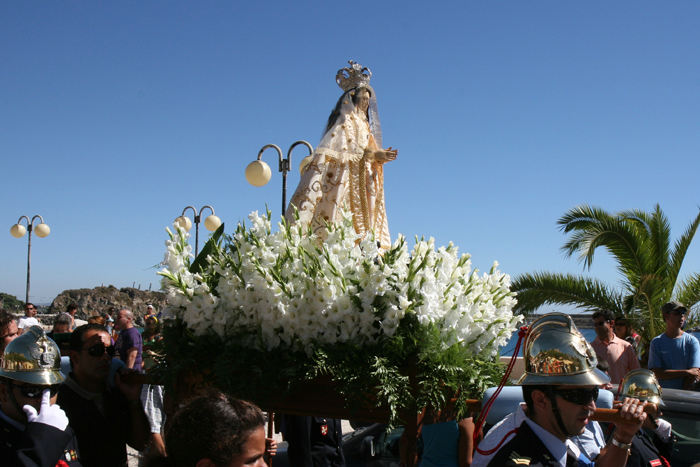 Nossa Senhora das Salas bæres rundt af 4 mand på en ’båre’ pyntet med blomster i dagens anledning. Hun er flankeret af byens spidser i galla, her skimtes brandmajoren.